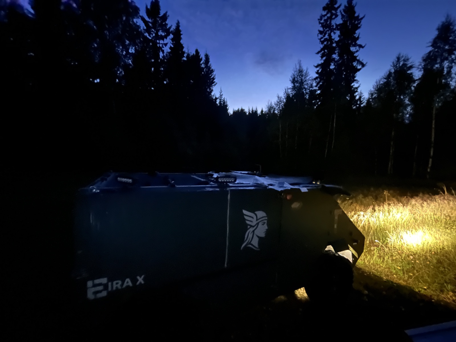 Eira X at dusk at the edge of a forest, its work lights on the grass