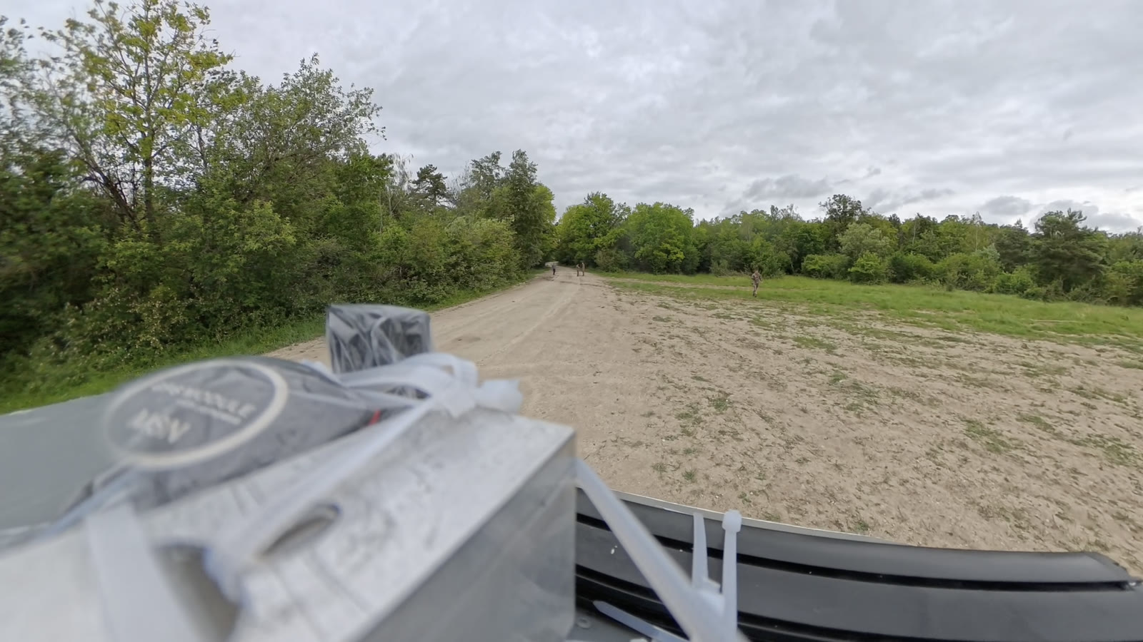 Camera view from the vehicle across a sandy track towards soldiers at the tree line